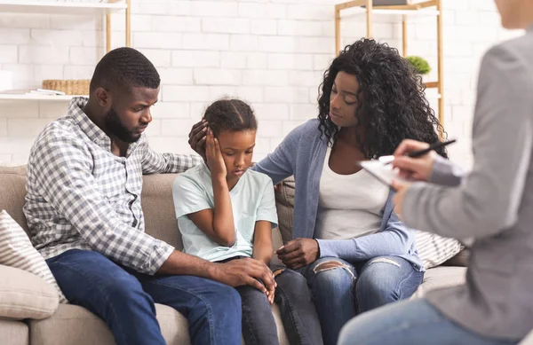 Little african american girl and her parents at reception of psychologist, kid sharing her feelings with doctor