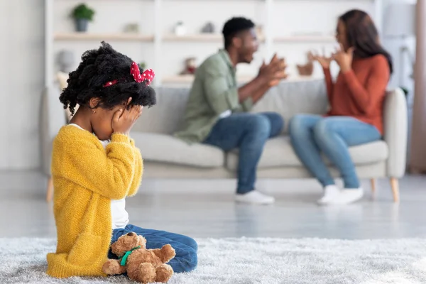 Kids And Domestic Conflicts. Little African American Girl Crying While Her Parents Arguing At Home, Upset Black Female Child Sitting On Floor With Teddy Bear And Covering Face, Selective Focus
